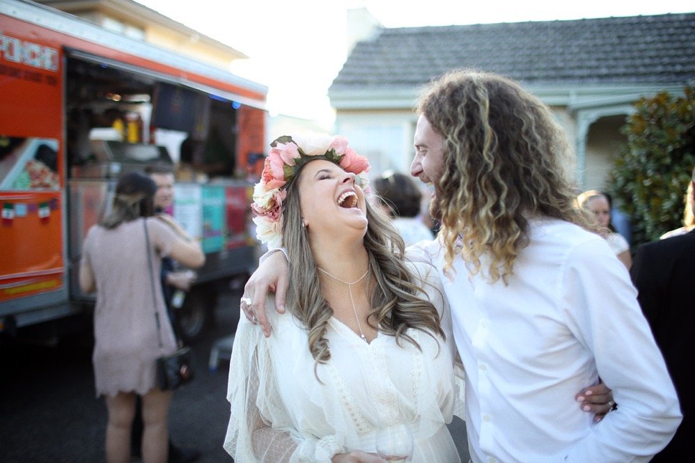 Bride in a flower crown and veil laughs with a long-haired man beside a food truck.