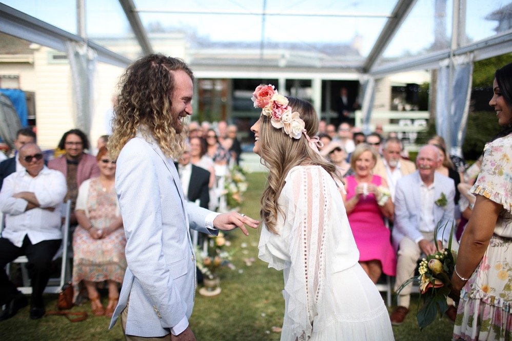 Bride in a white dress and flower crown faces groom in a light suit as guests watch under a clear tent.