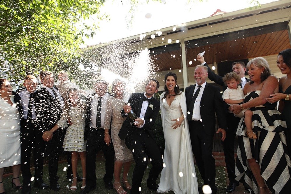 Bride and groom with guests as champagne sprays during an outdoor wedding celebration.