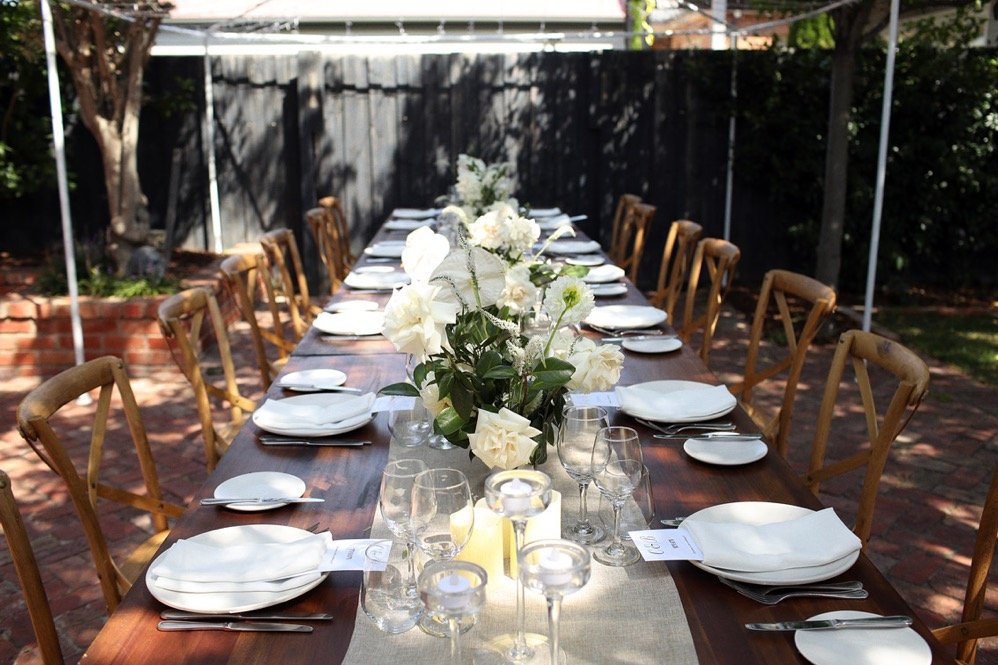 Outdoor wooden table set with white plates, glassware, candles and white floral centerpieces beneath a canopy.