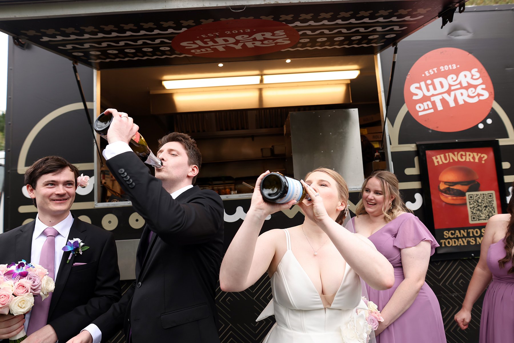 Bride and groom drink from bottles beside bridesmaids in front of a food truck.
