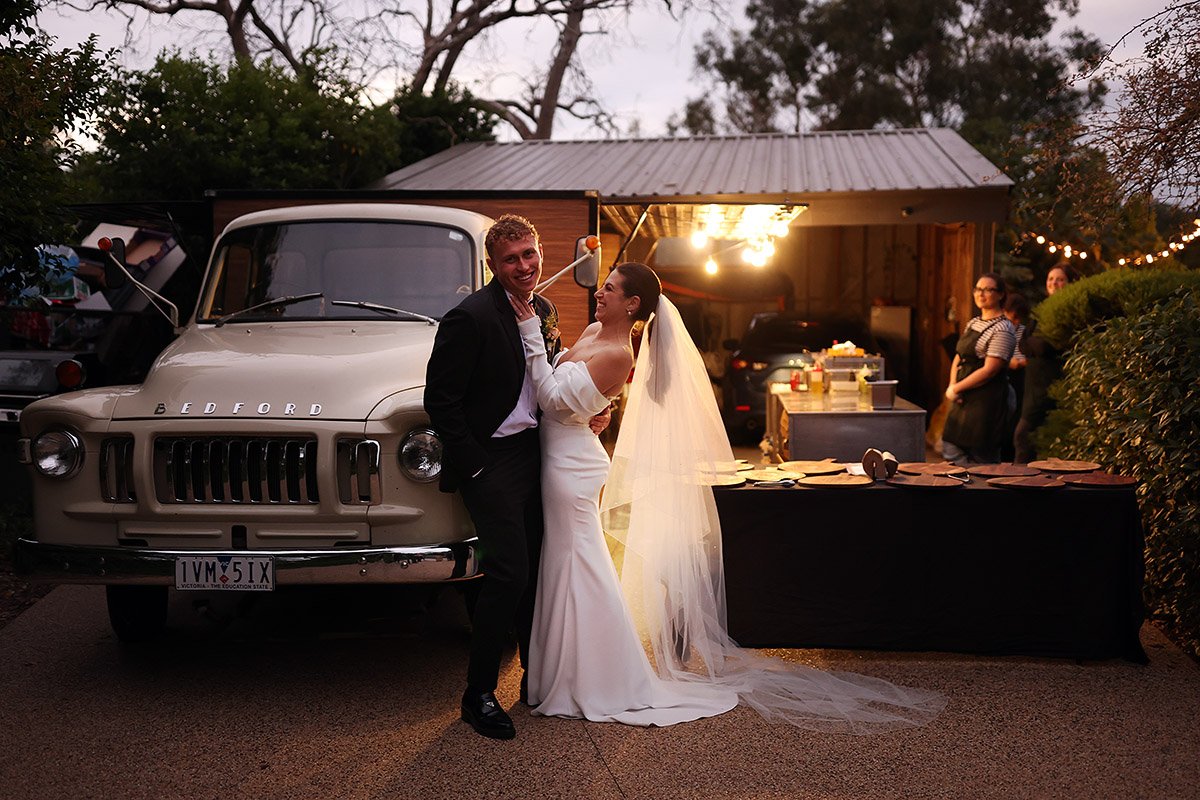 Bride and groom beside a vintage Bedford truck near a food table outside a wooden building.