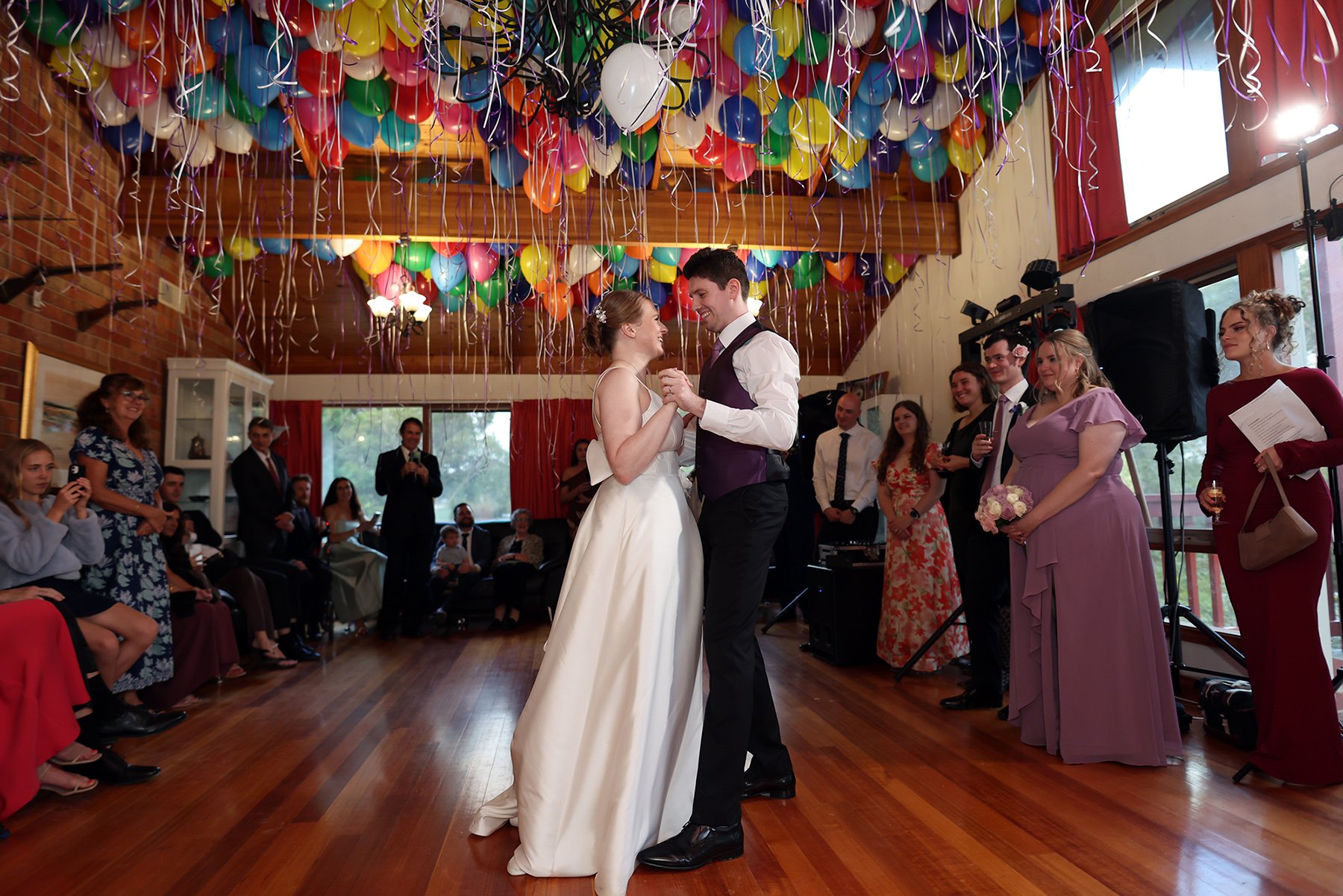 Bride and groom dance beneath colorful balloons and ribbons as wedding guests watch in a hall.