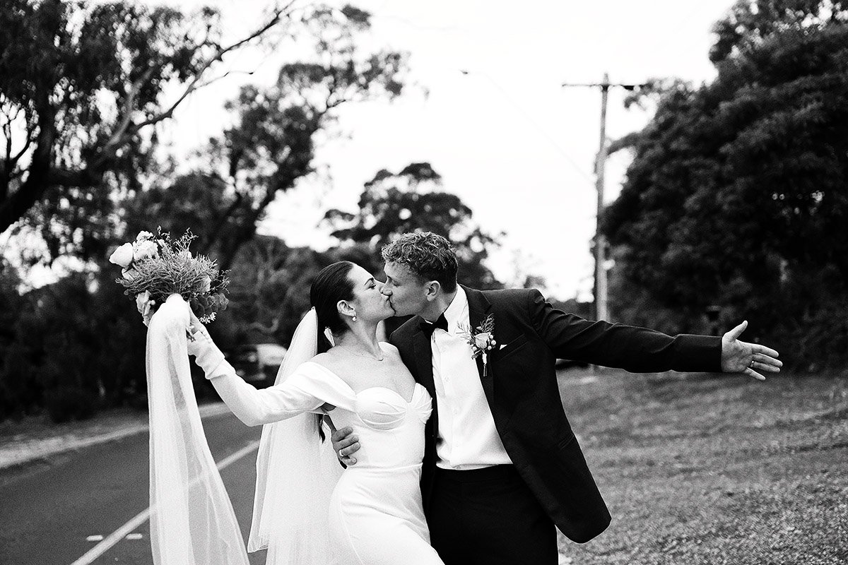 Bride and groom kissing on a tree-lined road, with bouquet and veil visible.
