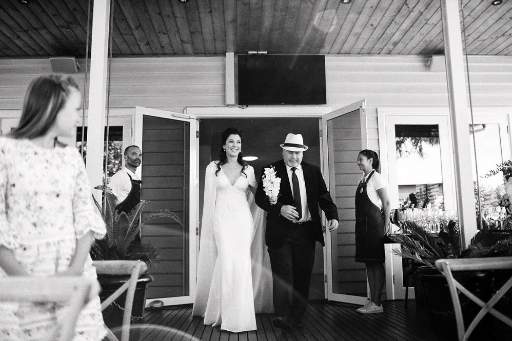 Bride and suited man walk through open doors onto a covered wooden porch with guests nearby.