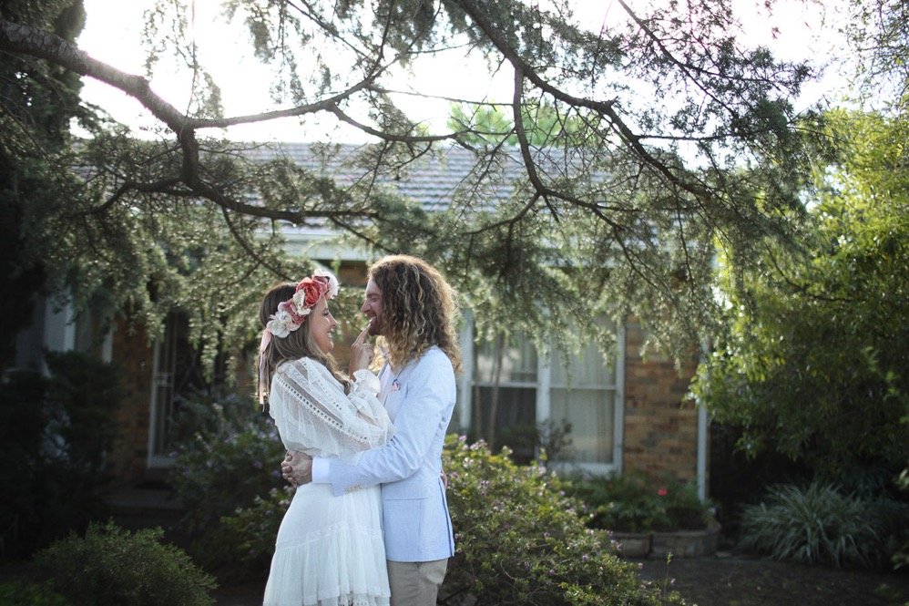 Bride in a white dress and groom in a light jacket standing together in a garden before a house.