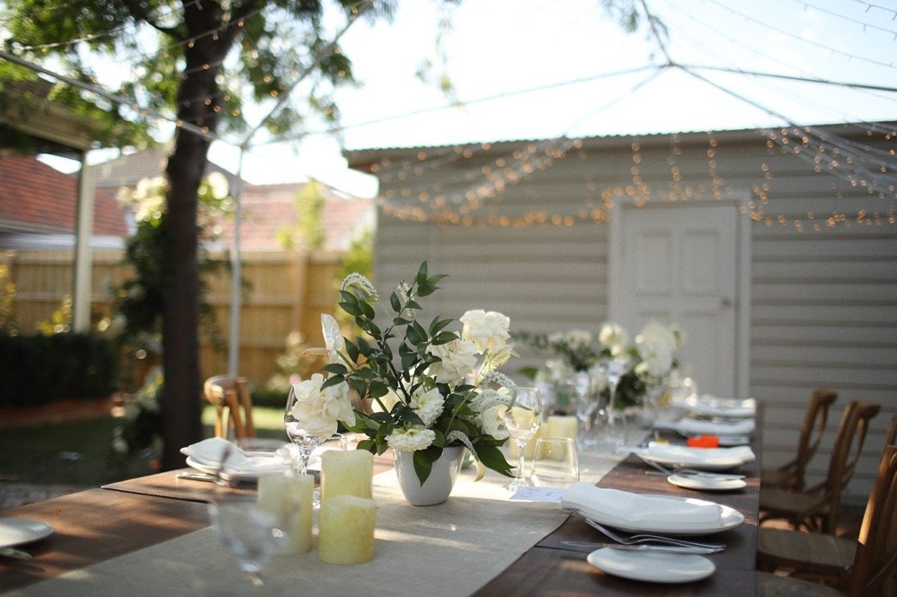 Outdoor wedding reception table with white flowers, candles, place settings, wooden chairs, and string lights.