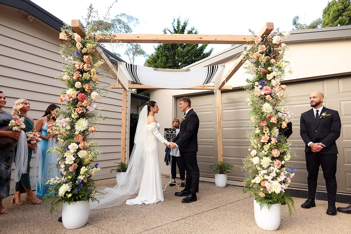 Bride and groom hold hands beneath a floral wooden canopy as attendants stand nearby.