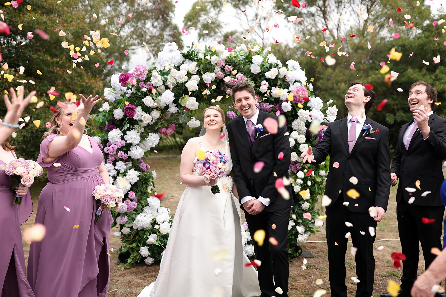 Bride and groom beneath a flower-covered arch as attendants toss flower petals.